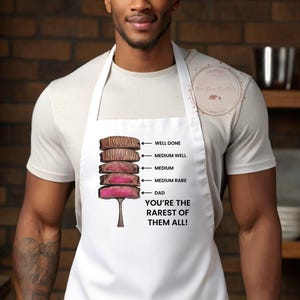 Könnte beinhalten: Weißer Schürze mit einer Grafik von Steak-Cuts, beschriftet von "Well Done" bis "Medium Rare", mit dem Text "DAD YOU'RE THE RAREST OF THEM ALL!". Die Schürze wird von einer Person getragen.