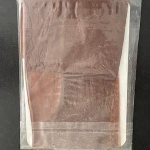 Puede incluir: Una tableta de chocolate rectangular de color marrón, envuelta en una funda de plástico transparente. El chocolate tiene una superficie lisa y plana, y está colocado sobre una tarjeta blanca visible a través del envoltorio protector.