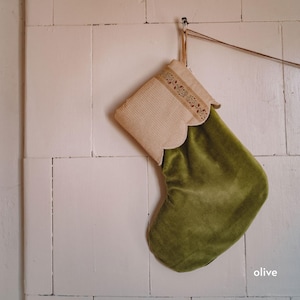 Op de afbeelding: Een olijfgroene kerstkous van fluweel met een beige boord met een subtiele krijtstreeptextuur en een geschulpte rand. De boord is versierd met een dunne, horizontale band van geborduurde bloemmotieven in gedempte rode en groene tinten.