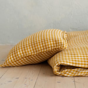 Puede incluir: Una almohada y ropa de cama a juego de vichy amarillo y blanco. La almohada está doblada y la ropa de cama está doblada sobre una superficie de madera.