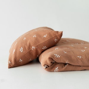 May include: A brown and white patterned muslin blanket and pillow set. The blanket and pillow are folded and placed on a white surface.