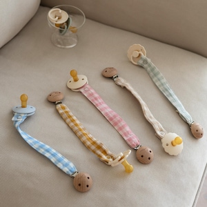 Puede incluir: Una colección de clips para chupetes de bebé en varios colores pastel, incluyendo azul, amarillo, rosa y beige. Cada clip presenta un botón de madera y un chupete a juego. Los chupetes están hechos de un material suave y natural.