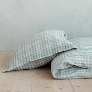 Pode incluir: Duas almofadas xadrez verdes e brancas e um cobertor dobrado repousam sobre um chão de madeira clara. O fundo é uma parede texturizada azul clara. A roupa de cama tem uma aparência macia e convidativa.
