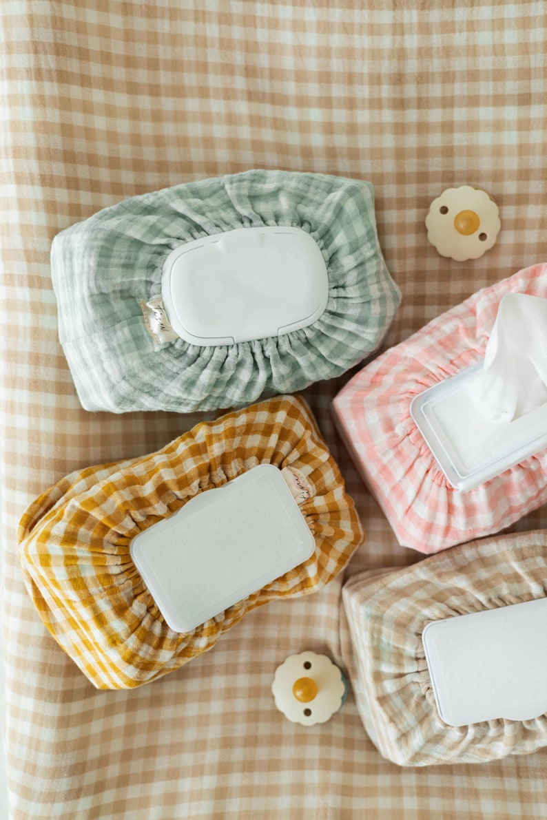 Op de afbeelding: Vier verschillende gepatroneerde stoffen hoezen voor babydoekjescontainers. De hoezen zijn in groen, geel, roze en beige. Elke hoes heeft een wit plastic deksel met een afgeronde rand.