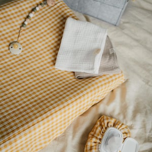 Op de afbeelding: Een gele en witte gingham verschoningsmat met een houten kralenspeelgoed en een speen. Er zijn twee witte muslin doeken en een gingham doekjesdoos met een wit deksel.