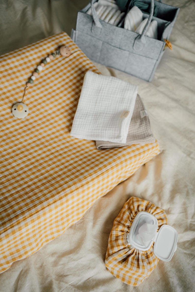 Op de afbeelding: Een gele en witte gingham verschoningsmat met een houten kralenspeelgoed en een speen. Er zijn twee witte muslin doeken en een gingham doekjesdoos met een wit deksel.