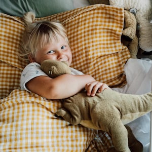 May include: A plush olive green dinosaur toy with a textured surface and visible stitched details, held by a child resting in a bed with yellow and white gingham patterned bedding and a solid sage green pillow.