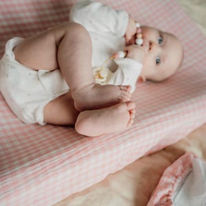 Op de afbeelding: Een baby die op een roze en witte geruite verschoningmat ligt, een wit rompertje draagt en een houten bijtring vasthoudt.
