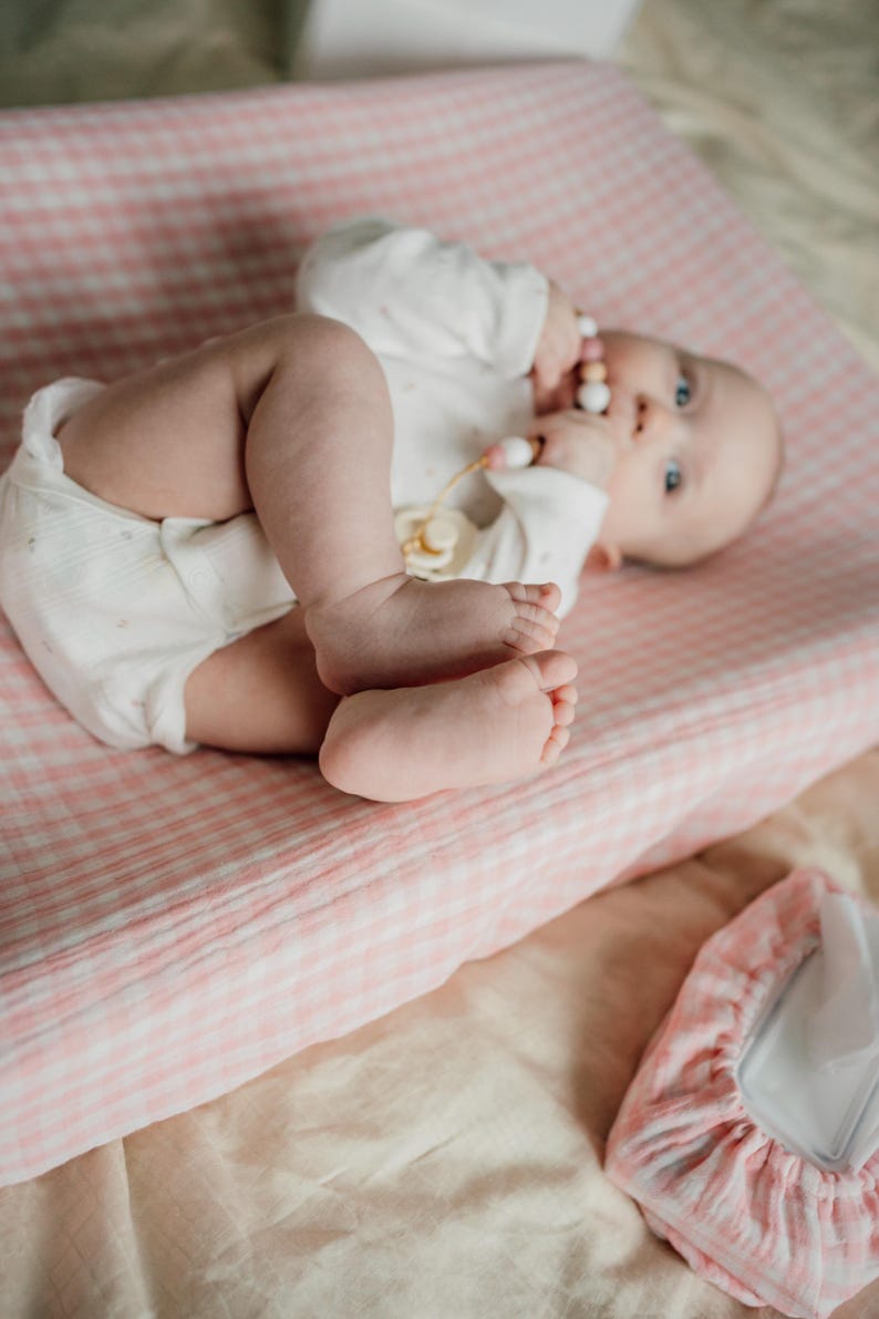 Op de afbeelding: Een baby die op een roze en witte geruite verschoningmat ligt, een wit rompertje draagt en een houten bijtring vasthoudt.