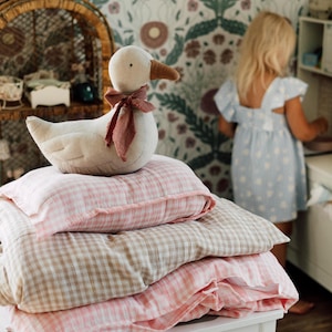 Pode incluir: Um brinquedo de pelúcia em forma de ganso com bico castanho e uma fita rosa-escura atada ao pescoço, pousado sobre uma pilha de três almofadas retangulares. As almofadas apresentam um tecido macio e texturado com um padrão xadrez rosa e bege.