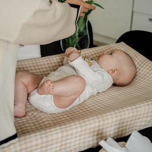 Op de afbeelding: Een baby ligt op een beige en bruine geruite verschoningmat. De baby draagt een wit rompertje met kleine bloemenmotieven. Een persoon houdt een boeket witte tulpen in een doorzichtige vaas.