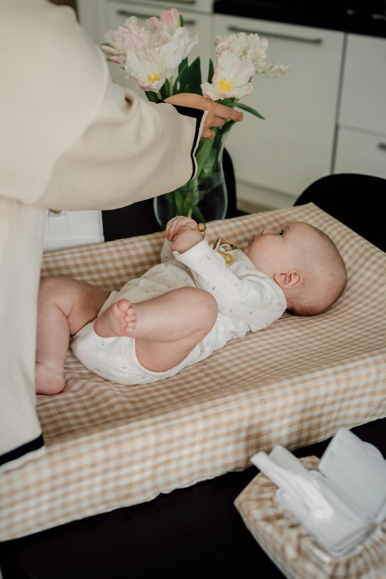 Op de afbeelding: Een baby ligt op een beige en bruine geruite verschoningmat. De baby draagt een wit rompertje met kleine bloemenmotieven. Een persoon houdt een boeket witte tulpen in een doorzichtige vaas.