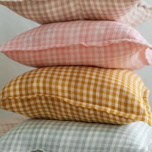Puede incluir: Una pila de cuatro almohadas de gingham en diferentes colores. Las almohadas son beige, rosa, amarillo y verde. Las almohadas están apiladas una encima de la otra, con la almohada beige en la parte superior.