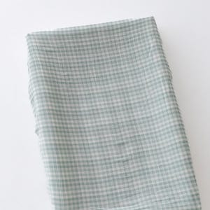 Könnte beinhalten: Ein grüner und weißer Gingham-Stoff mit weicher, strukturierter Haptik. Der Stoff ist gefaltet und liegt flach auf einer weißen Oberfläche.