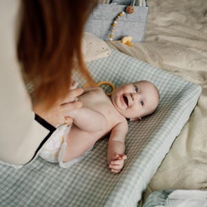 Op de afbeelding: Een baby ligt op een groene en witte geruite verschoningsmat, glimlacht en kijkt omhoog. De baby draagt een luier en heeft een houten ring in zijn hand. De verschoningsmat ligt op een beige oppervlak.