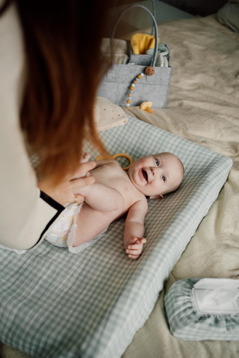 Op de afbeelding: Een baby ligt op een groene en witte geruite verschoningsmat, glimlacht en kijkt omhoog. De baby draagt een luier en heeft een houten ring in zijn hand. De verschoningsmat ligt op een beige oppervlak.