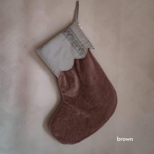 Könnte beinhalten: Ein brauner Weihnachtsstrumpf aus Samt mit einer hellgrauen Manschette mit gewelltem Rand und einem feinen vertikalen Streifenmuster. Die Manschette ist mit einem Mittelband verziert, das ein kleines Blumenmuster in gedeckten Rot- und Grüntönen zeigt.