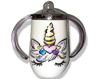 May include: White stainless steel sippy cup with a unicorn design. The unicorn has a gold horn, pink and blue hearts, and purple and pink mane. The cup has two clear plastic handles.
