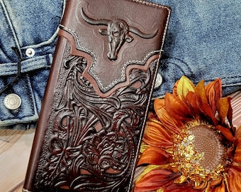 May include: A brown leather wallet with a tooled design featuring a bull's head and floral patterns. The wallet is closed and lying on a denim background with a brown sunflower.