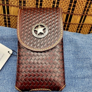 May include: A brown leather phone case with a woven pattern and a silver star emblem. The case has a flap closure and yellow stitching around the edges. The case is designed to hold a phone.