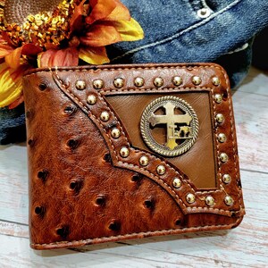May include: A brown leather wallet with a cross design and gold accents. The wallet has a textured, ostrich-like pattern and is decorated with gold studs.