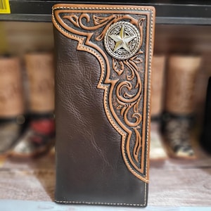 May include: A brown leather wallet with a decorative embossed design and a silver star emblem. The wallet features a contrasting tan border and stitching. The design has a western theme.