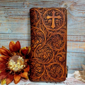 May include: A brown leather wallet with a floral and cross design. The wallet has a zipper closure and is decorated with embossed flowers and a cross. The wallet is a long, rectangular shape.