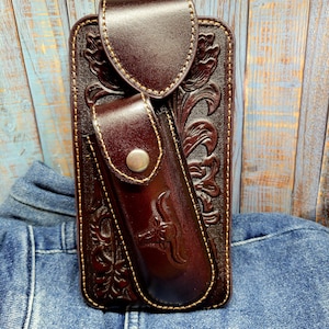 May include: A dark brown leather phone case with a matching belt clip. The case features intricate floral and scrollwork designs, with a smaller pouch attached. The pouch has a silver button closure and a bull graphic.