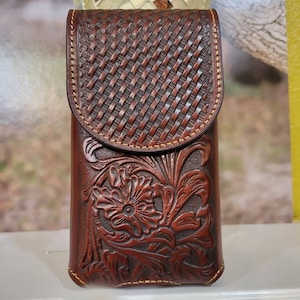 May include: A brown leather phone case with a floral design and a woven pattern on the flap. The case is stitched with tan thread and has a snap closure. The case is approximately 6 inches tall.