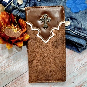 May include: A brown leather wallet with a white and brown floral embossed design. The wallet has a metal cross design on the front.