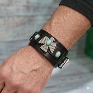 May include: Black leather cuff bracelet featuring a silver-tone Thunderbird emblem and three turquoise-colored stones. The bracelet has a buckle closure and is worn on a wrist.