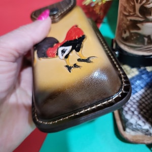 May include: A handcrafted leather wallet with a rooster design. The wallet features a tan and brown gradient, with a detailed rooster illustration in red, black, and yellow. The wallet has a stitched border and a small flap.