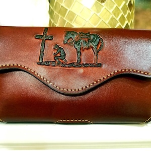May include: A brown leather wallet with a flap closure. The front features a detailed embossed design of a cross, a kneeling figure, and a horse. The edges are stitched with a contrasting thread.