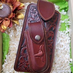 May include: A set of handcrafted, dark brown leather knife sheaths with intricate floral and animal carvings. The set includes a large sheath, a smaller sheath, and a belt loop, all stitched with contrasting thread. The leather has a rich, textured appearance.