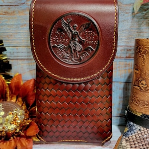 May include: A handcrafted, dark brown leather phone case with a woven pattern on the front. The flap features a detailed embossed design of a coin with an angel. The case is stitched with a contrasting light brown thread.