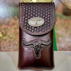 May include: A brown leather phone case with a woven pattern and a silver metal bull head design. The case has a flap with a silver metal buckle with a bull head design.