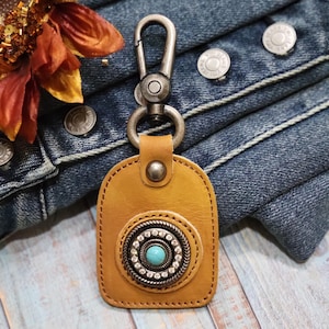May include: A tan leather keychain with a turquoise stone and silver accents. The keychain has a metal clasp and a loop for attaching keys.