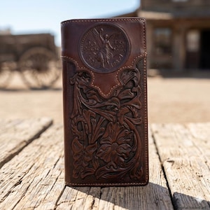 May include: A brown leather wallet with intricate floral and leaf tooling. The wallet features a circular emblem with text and a figure. The design is embossed on the front of the wallet.