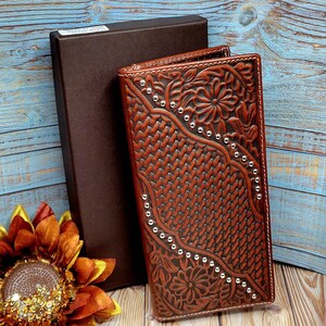 May include: A brown leather wallet with a floral and woven design, accented with silver studs. The wallet is next to a dark brown box. The background is a blue wooden surface. The wallet is a long, rectangular shape.