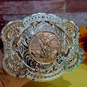May include: Ornate silver and gold-tone belt buckle featuring a central gold coin with a winged figure and the text "50 Pesos." The buckle has detailed floral and feather designs. A decorative accessory.