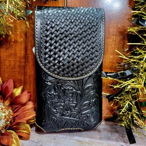May include: Black leather phone case with a woven top flap and floral embossed design. The case has a curved top and is stitched with a light-colored thread. The case is rectangular and has a snap closure.