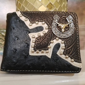 May include: Black leather wallet with a white and brown embossed design. The wallet features a silver horseshoe with a steer head and studded accents.