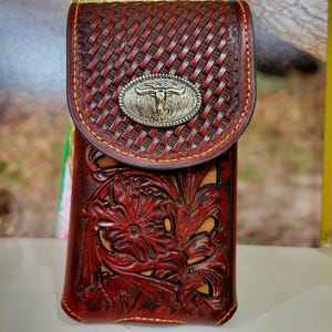 May include: A red leather phone case with a woven pattern and a silver metal buckle with a steer head design. The case has a floral design embossed on the front.
