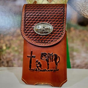 May include: A brown leather phone case with a decorative flap. The flap has a woven pattern and a silver-colored oval buckle. The front of the case features a cross, a kneeling figure, and a horse.