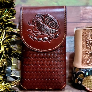 May include: A brown leather phone case with a woven pattern and a flap. The flap features an embossed design of an eagle with a snake in its beak, a symbol of Mexico. The case is stitched with a contrasting thread.