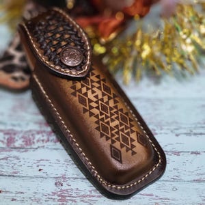 May include: A brown leather knife sheath with a geometric pattern and a decorative button. The sheath has a stitched border and a textured top flap. The design is embossed into the leather.