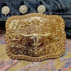 May include: A gold-colored rectangular belt buckle with a detailed design featuring a longhorn steer, horseshoe, and floral accents. The buckle is embellished with small, clear rhinestones around the edges and has a silver-colored steer head.