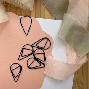 May include: A close-up of a set of black metal paper clips shaped like teardrops. The clips are arranged on a pink and white surface.