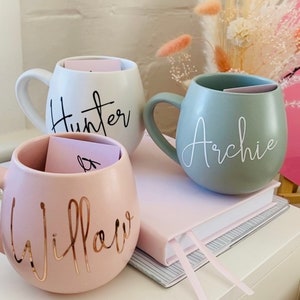 May include: Three ceramic mugs in white, sage green, and pink, each with a curved handle and a name in a cursive font. The white mug says "Hunter" in black, the sage green mug says "Archie" in white, and the pink mug says "Willow" in gold.