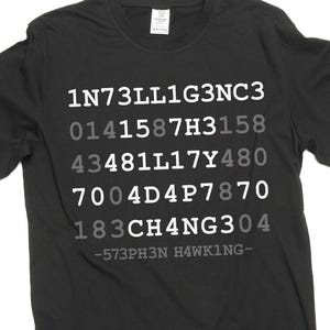 Op de afbeelding: Zwart t-shirt met witte en grijze tekst. De tekst bevat een reeks getallen en letters, met de naam "HAWKING" onderaan. Het shirt heeft een ronde hals.